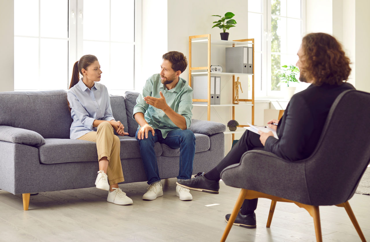 Young family couple sitting on sofa at therapist office, talking to each other about their relationship problems. Professional psychologist providing support to husband and wife during therapy session