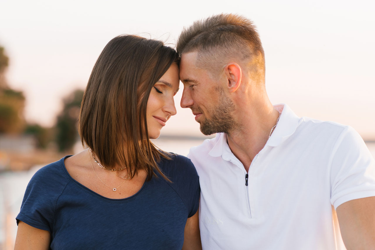 Loving couple standing close together at sunset by the water, looking into each other eyes with tenderness. Perfect for concepts of romance, intimacy, and relationships.